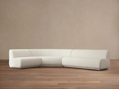 Gianni Three-Piece Sectional in off-white fabric, positioned on a wooden floor with a neutral beige background. Sectional features a curved left piece and long right piece.