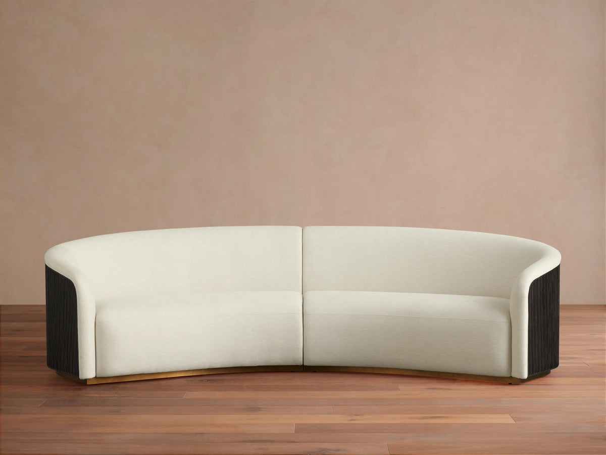 Finnley Two-Piece Curved Sectional in cream fabric, with dark fluted arm ends, and a golden base, on a wood floor.