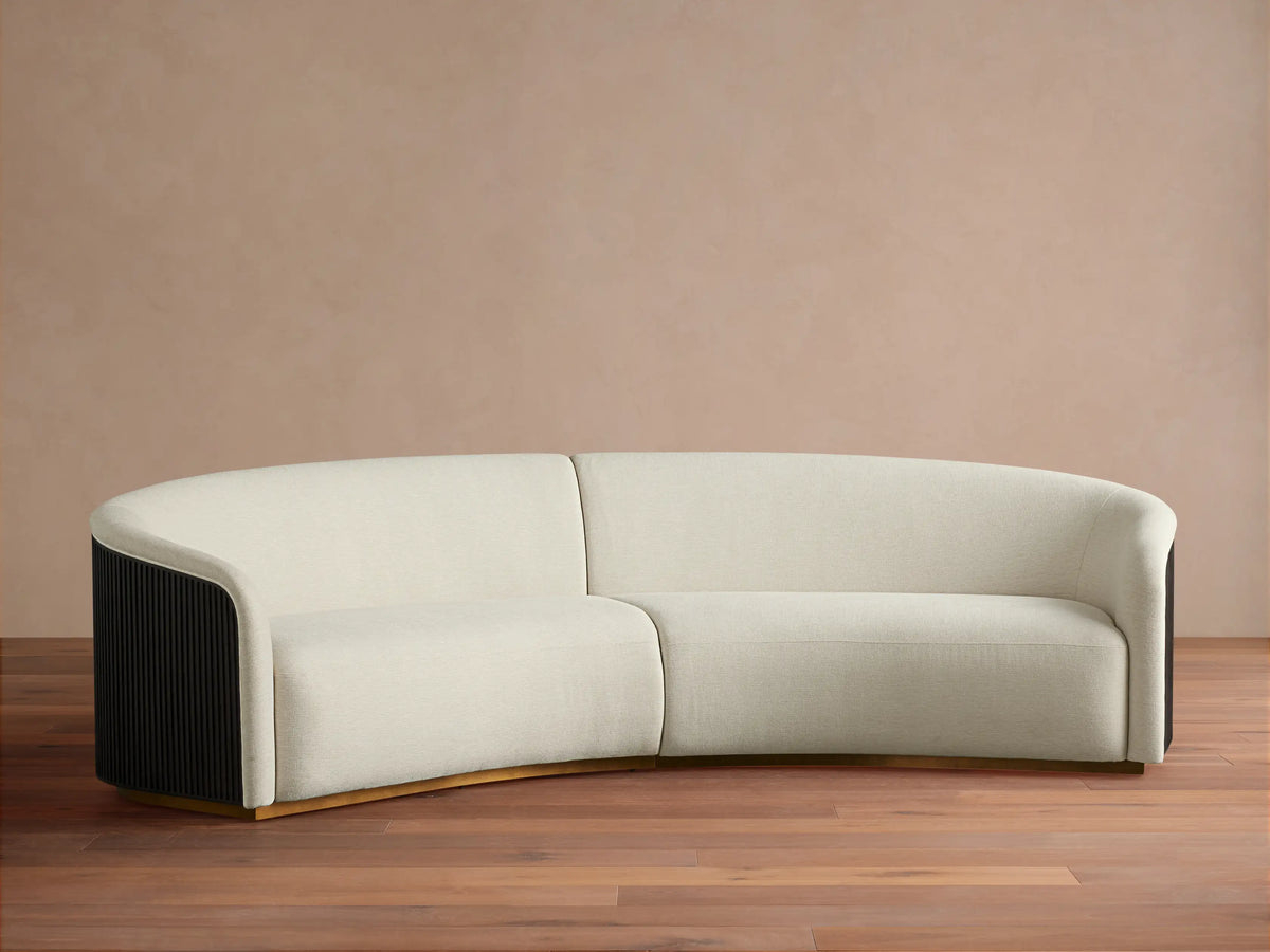 Finnley Two-Piece Curved Sectional in cream fabric, with dark fluted arm ends, and a golden base, on a wood floor.