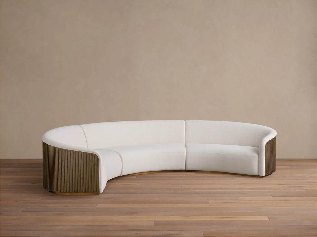 Curved three-piece sectional sofa, upholstered in soft white fabric, featuring wooden side panels and positioned on a wooden floor.