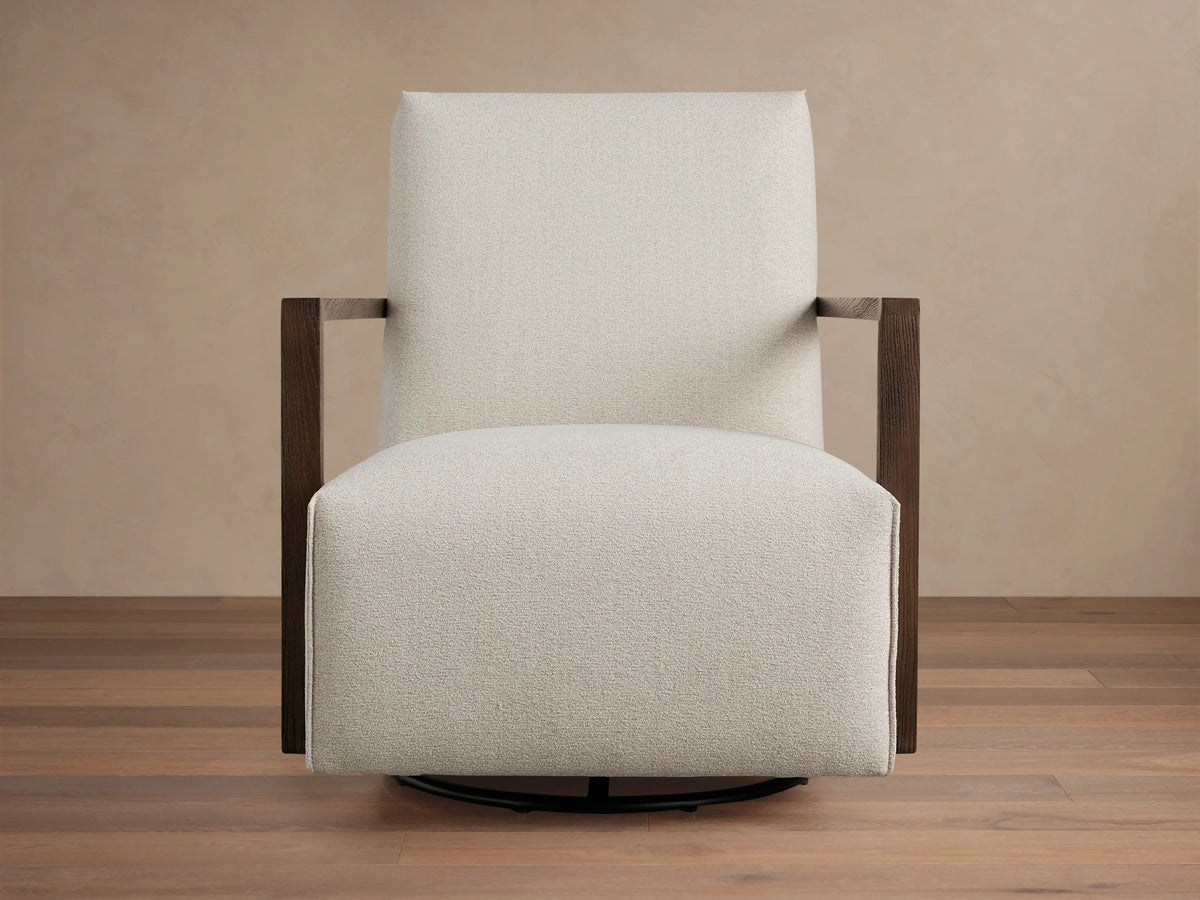 Walsh Glider in light beige fabric with square wooden arms on a round black swivel base, set against a wooden floor.