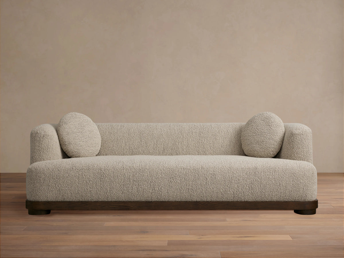 Marcela Sofa in beige upholstery against a neutral wall, featuring rounded armrests and two circular cushions, on a wooden floor.