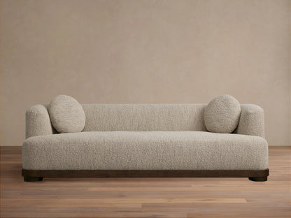 Marcela Sofa in beige upholstery against a neutral wall, featuring rounded armrests and two circular cushions, on a wooden floor.