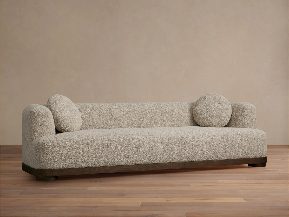 Marcela Sofa in beige upholstery against a neutral wall, featuring rounded armrests and two circular cushions, on a wooden floor.