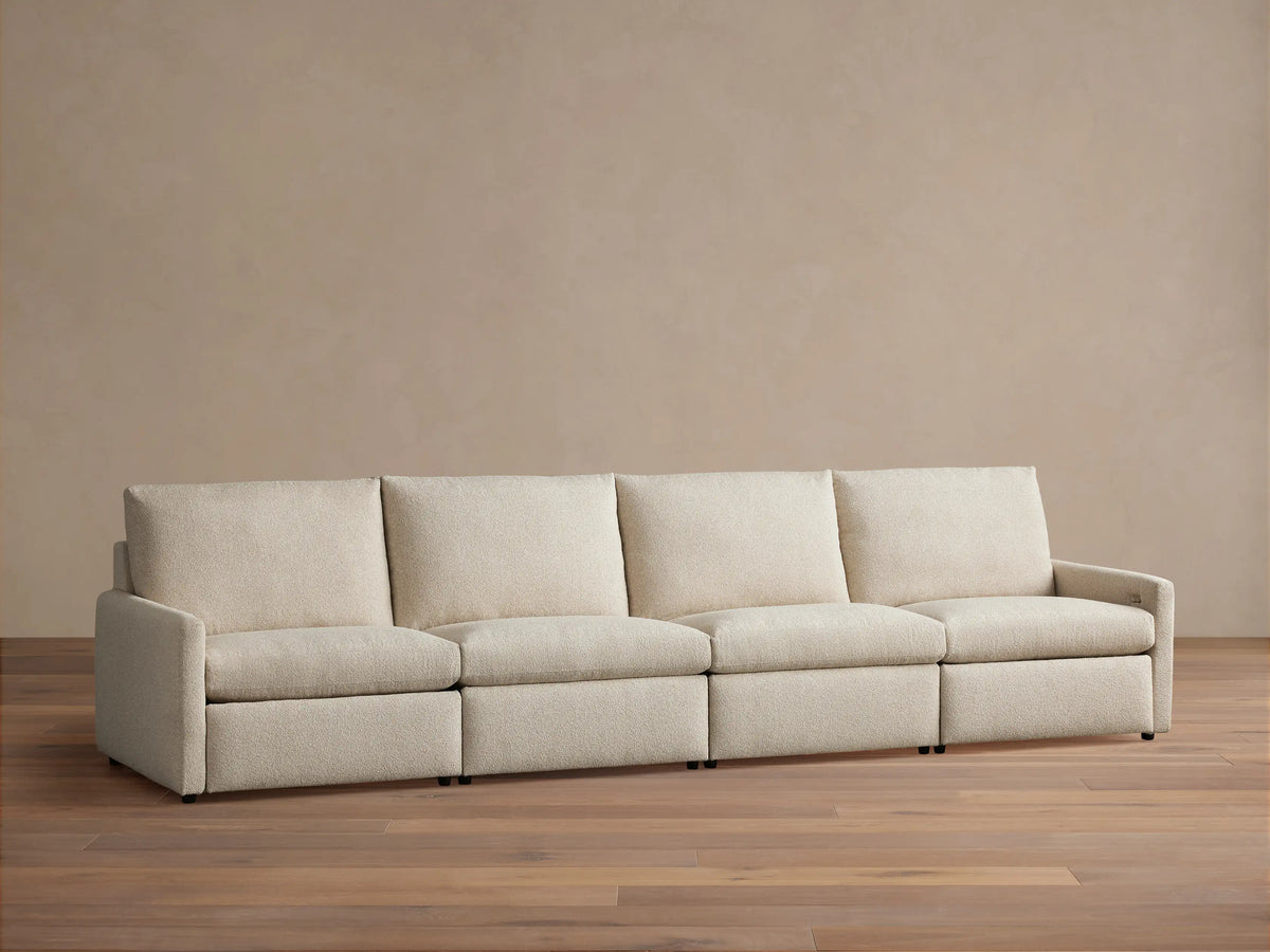 Kipton Four-Piece Motion Modular Sofa in beige fabric, viewed from the front. Features slim arms, four cushioned seats, and backrests, on a wooden floor.