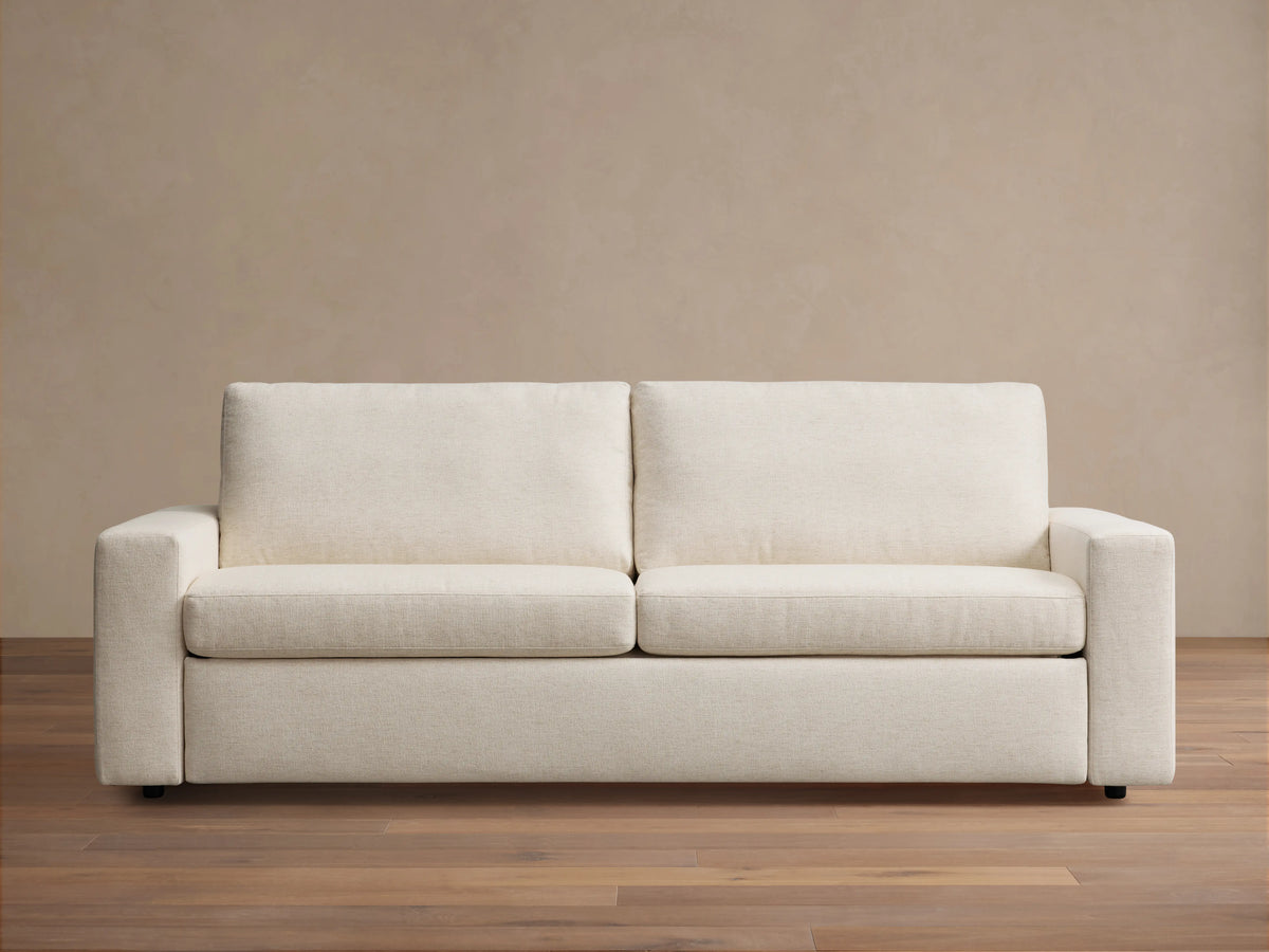 Kipton Wide Arm Luxury Sleeper Sofa in beige fabric, features wide arms, rectangular back cushions, and a sleek wood floor setting.