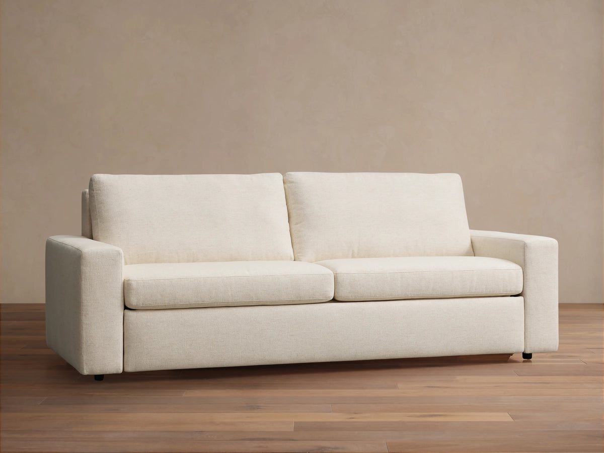 Beige Kipton Wide Arm Luxury Sleeper Sofa extended into a bed, showing white bedding and sturdy black legs against a light brown background.