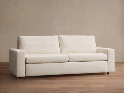 Beige Kipton Wide Arm Luxury Sleeper Sofa extended into a bed, showing white bedding and sturdy black legs against a light brown background.