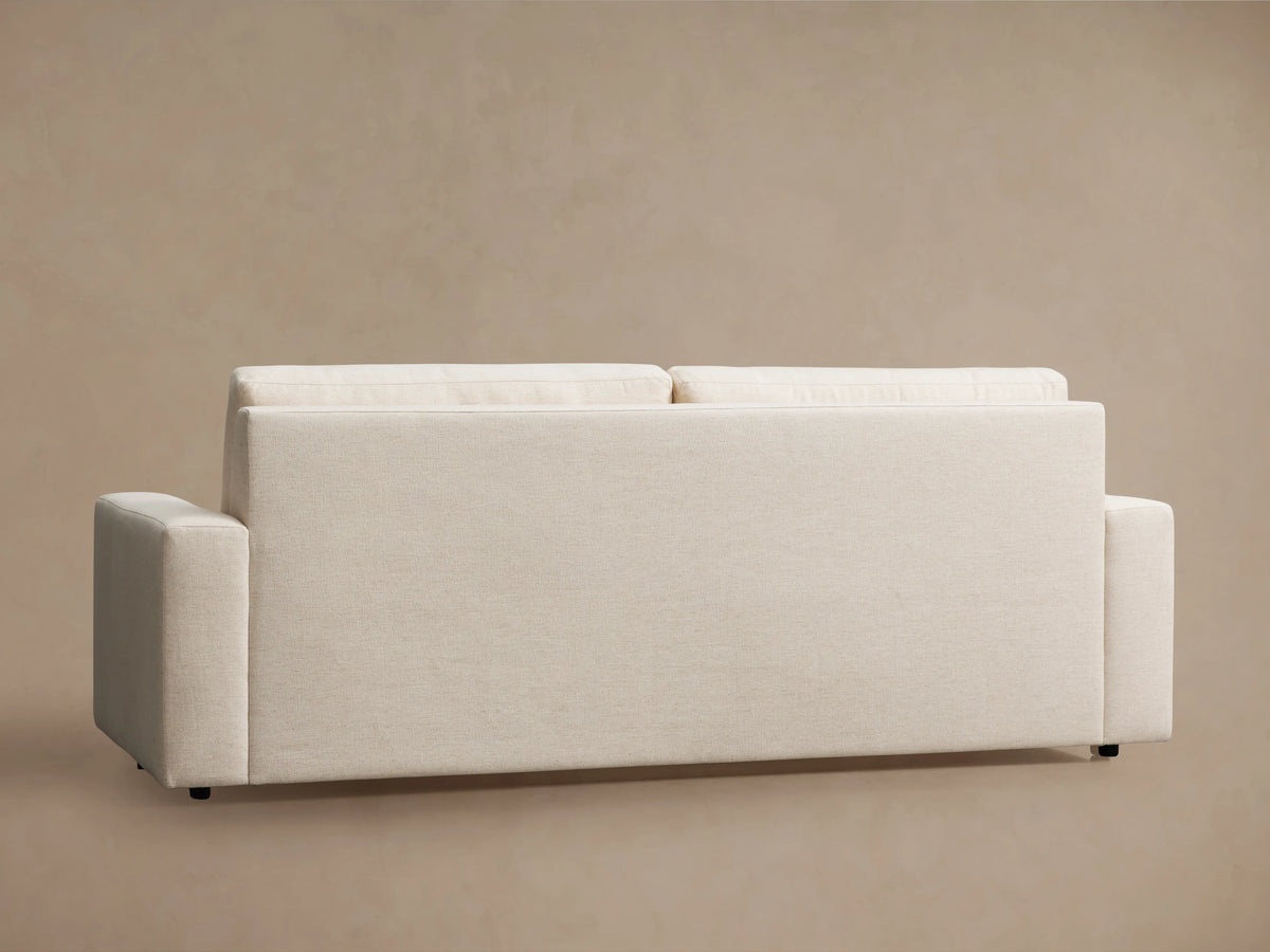 Kipton Wide Arm Luxury Sleeper Sofa in beige Crypton fabric, features wide arms, two seat cushions, and two back cushions, set against a light brown wall and wooden floor.