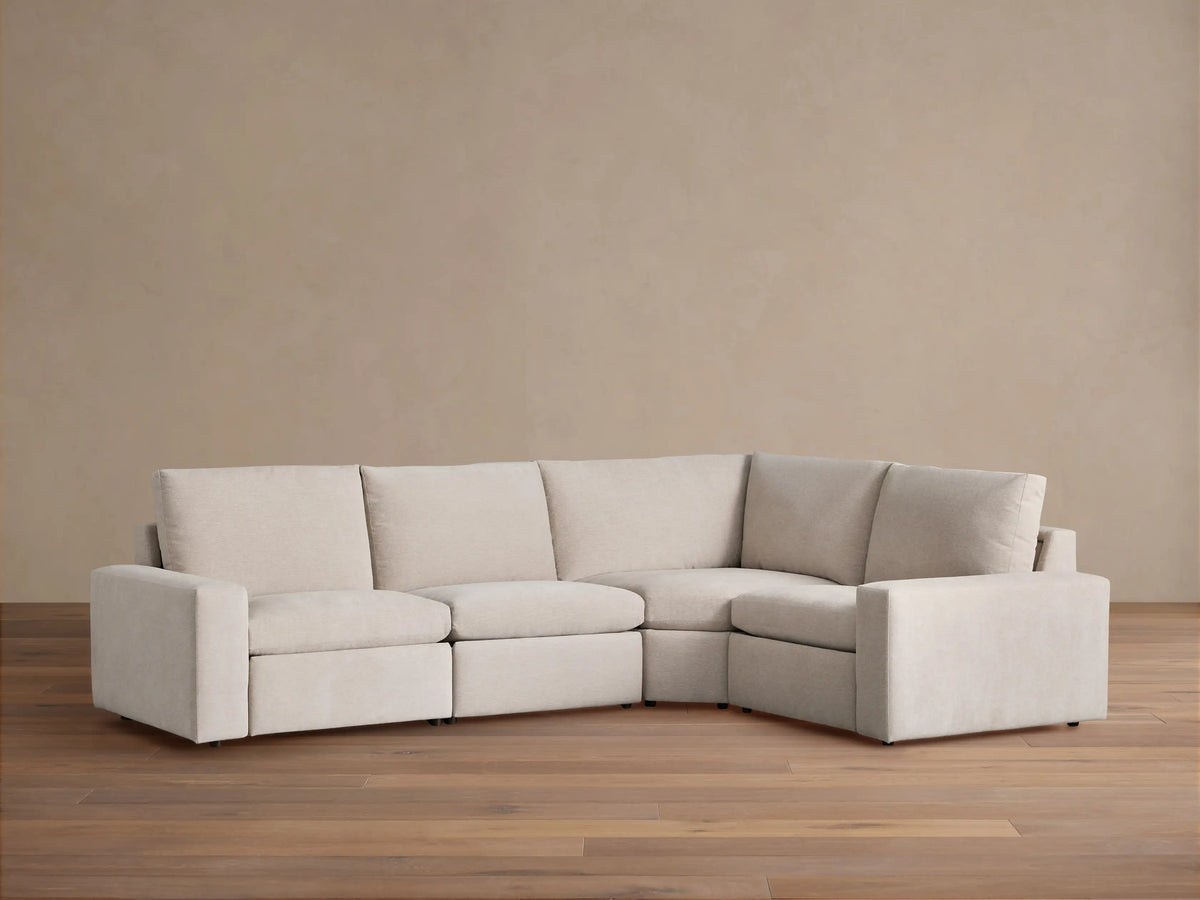 Kipton Wide Arm Four-Piece Motion Sectional in light beige fabric, arranged in an L-shape against a tan wall on a wood floor.