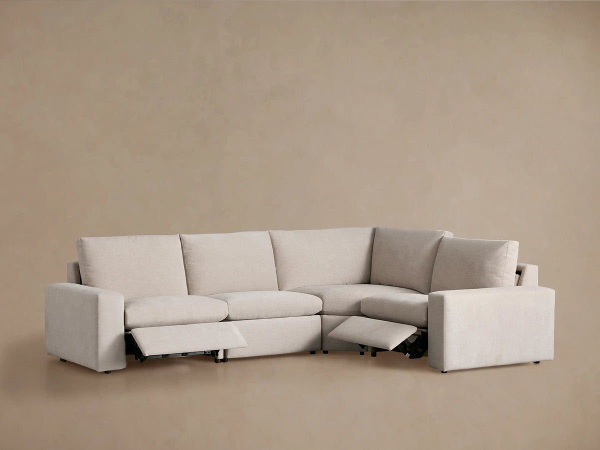 Kipton Wide Arm Four-Piece Motion Sectional in light beige textured fabric, seen from the back and side on a brown background.
