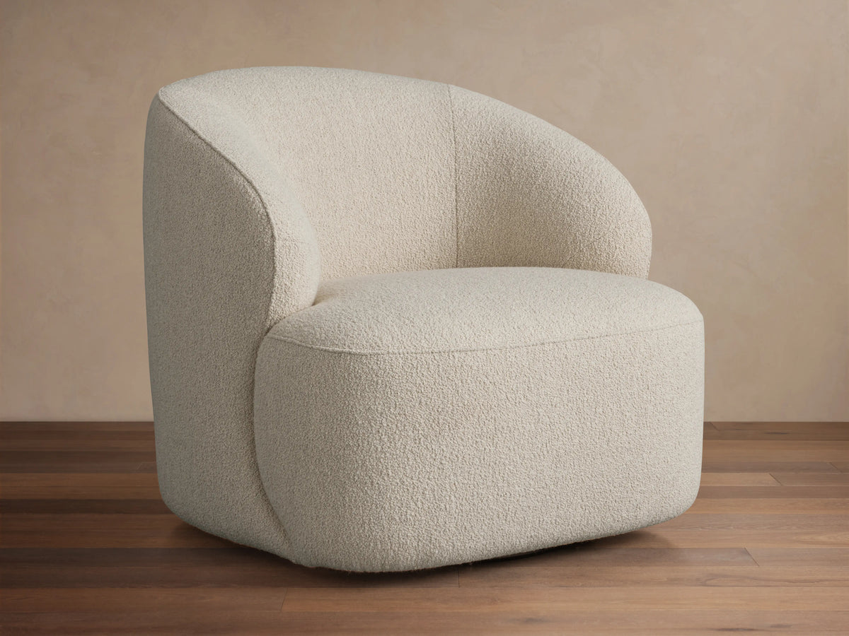 Alonzo Swivel Chair, cream bouclé, with a rounded backrest and seat, angled on a wooden floor against a beige wall.