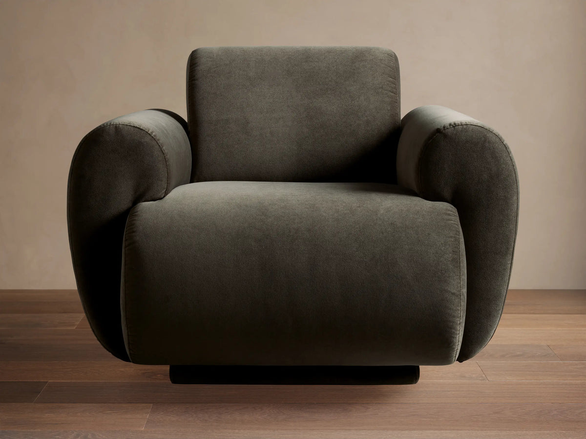 Gianni Swivel Chair in dark olive velvet, modern rounded design, angled slightly left, on a wooden floor against a beige wall.