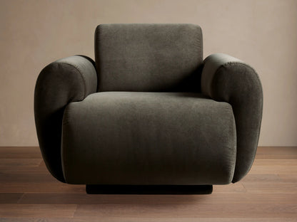 Gianni Swivel Chair in dark olive velvet, modern rounded design, angled slightly left, on a wooden floor against a beige wall.