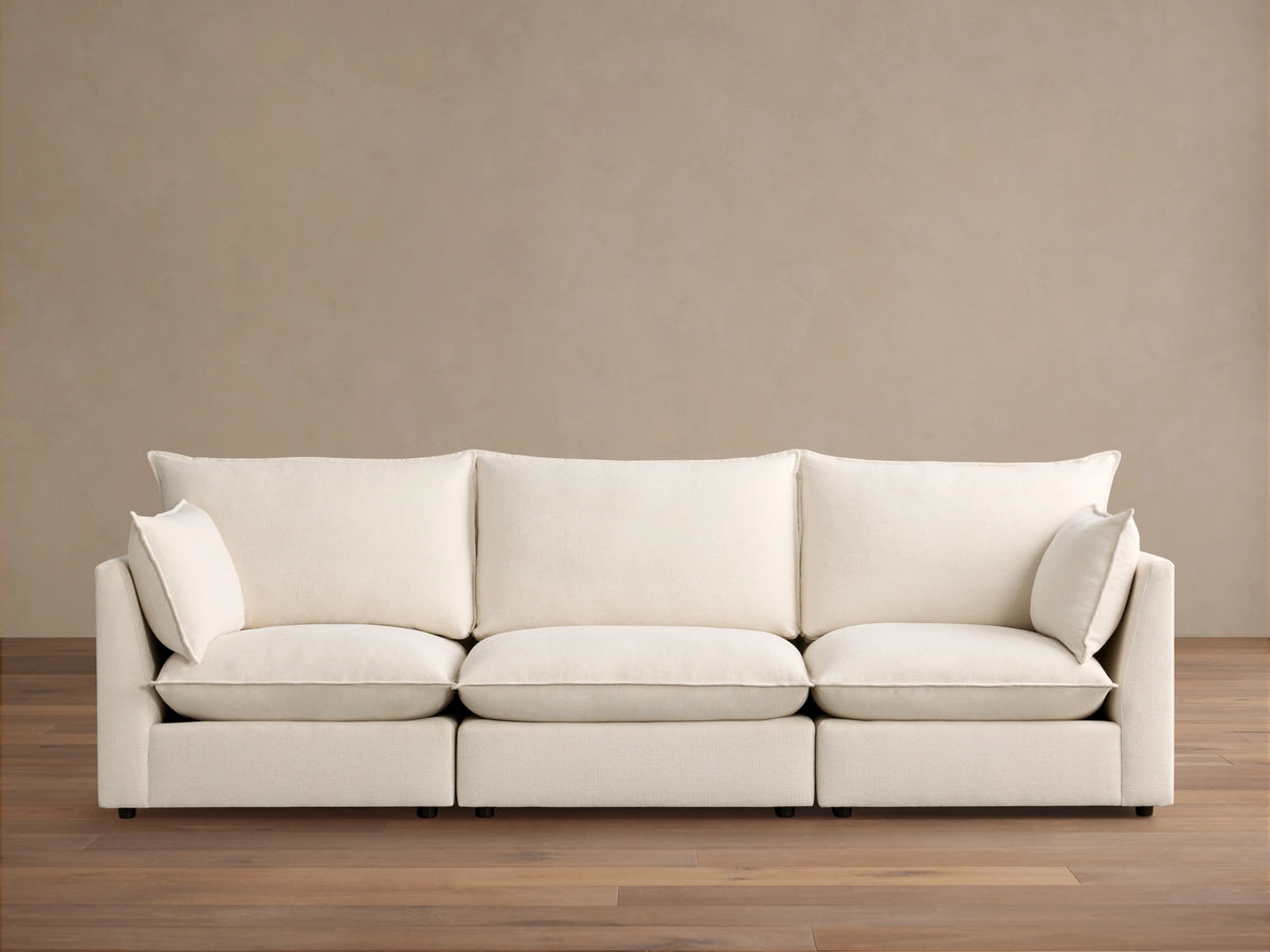 Owen Three Piece Modular Sofa in soft cream fabric, three seats with back cushions and armrests, on a wooden floor against beige wall.