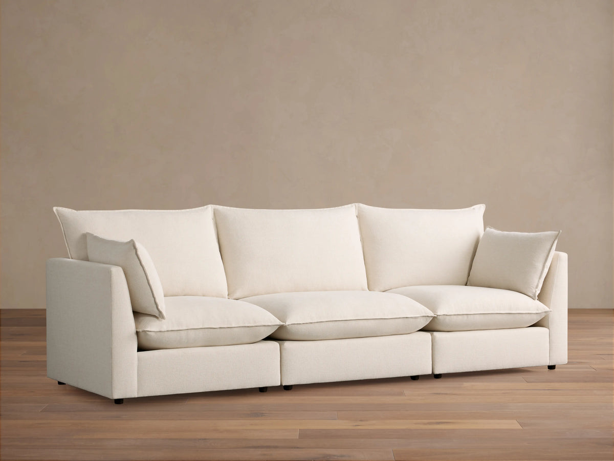 Owen Three Piece Modular Sofa in soft cream fabric, three seats with back cushions and armrests, on a wooden floor against beige wall.