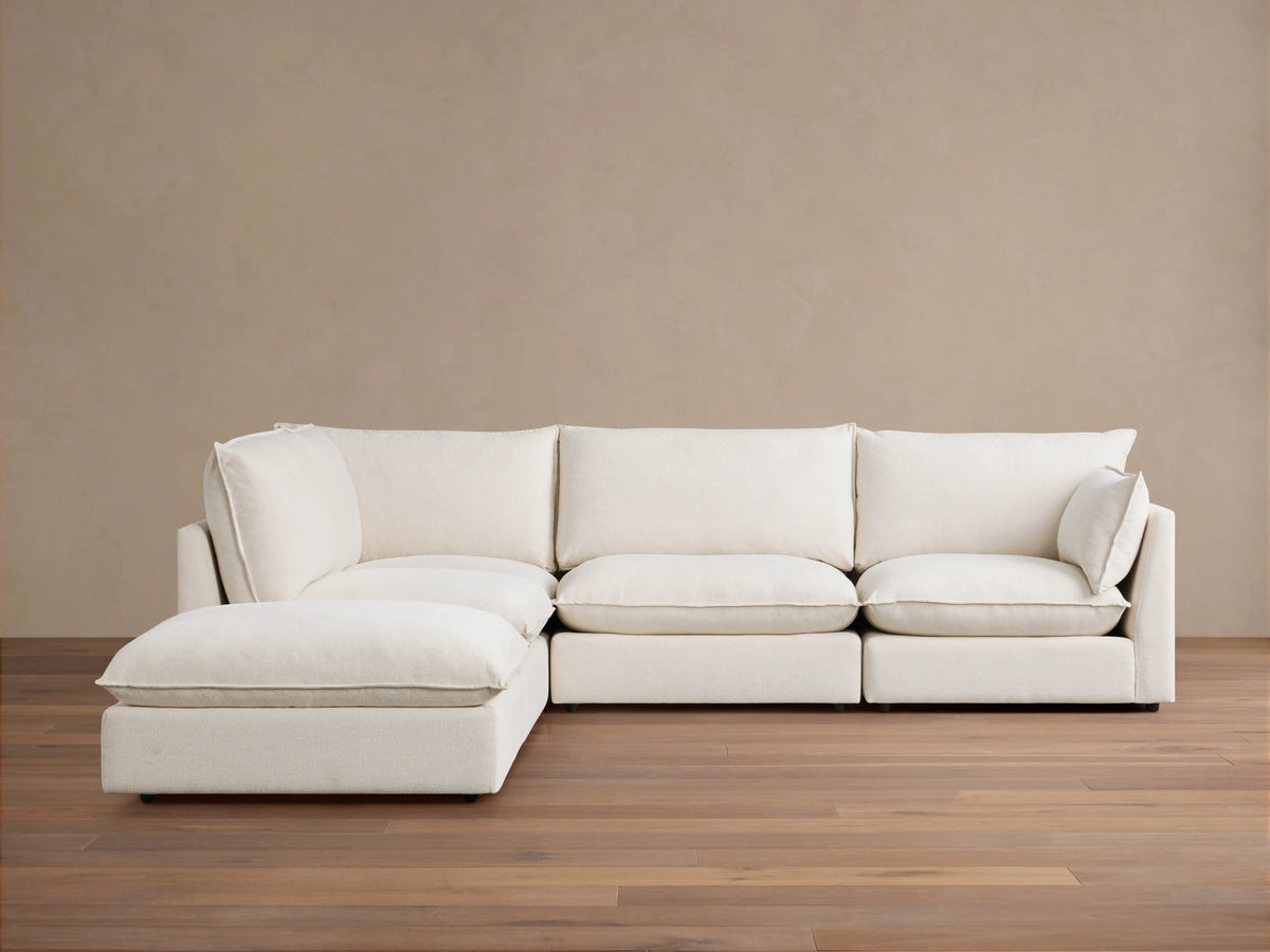 A cream-colored Owen Five-Piece Sectional in an L-shape placed on wooden flooring against a beige wall. The sectional has plush cushions and soft upholstery.