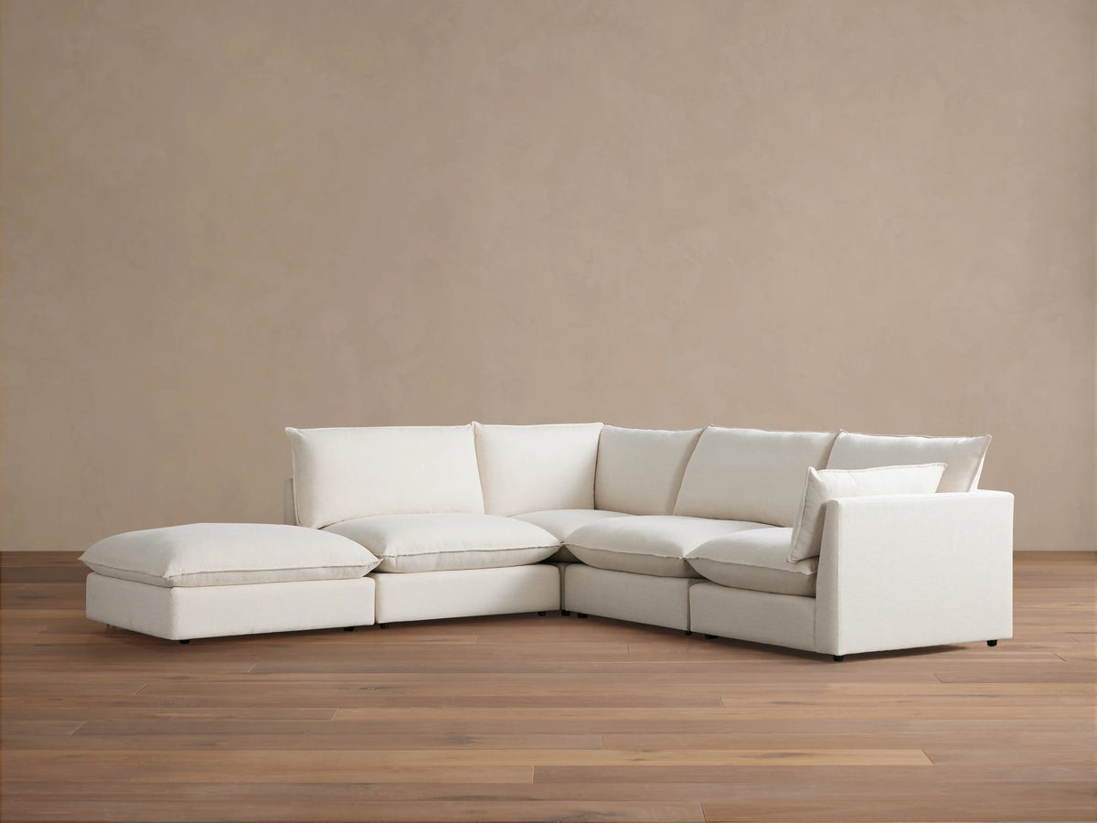 A cream-colored Owen Five-Piece Sectional in an L-shape placed on wooden flooring against a beige wall. The sectional has plush cushions and soft upholstery.