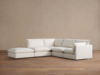 A cream-colored Owen Five-Piece Sectional in an L-shape placed on wooden flooring against a beige wall. The sectional has plush cushions and soft upholstery.