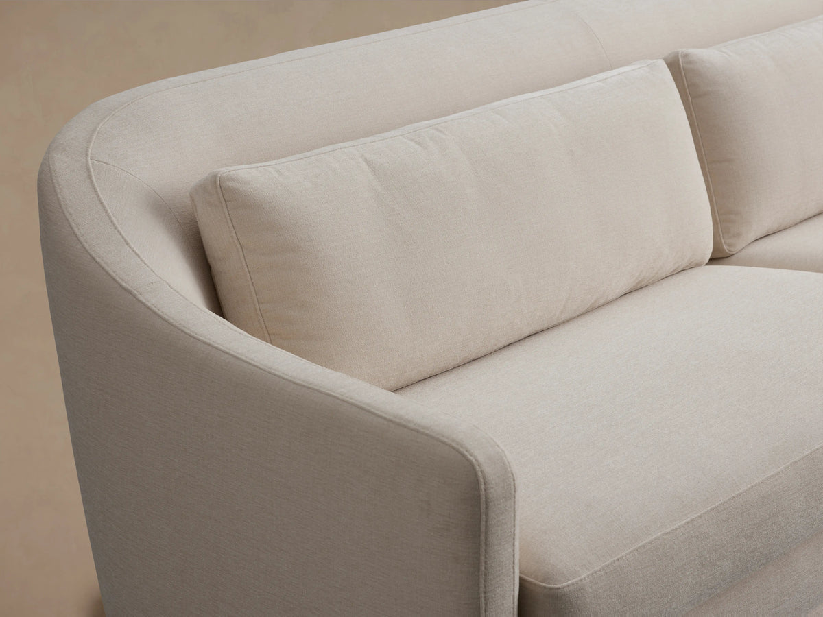 Bianca Sofa in light beige fabric, featuring a sleek, curved backrest with plush cushions. Positioned on a wooden floor, neutral backdrop.