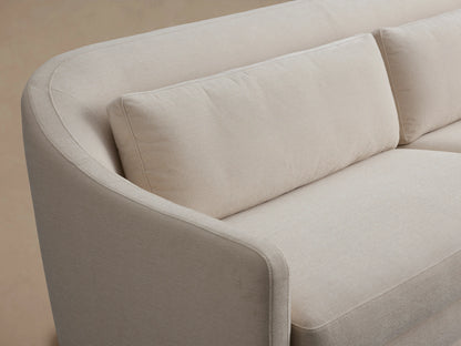 Bianca Sofa in light beige fabric, featuring a sleek, curved backrest with plush cushions. Positioned on a wooden floor, neutral backdrop.
