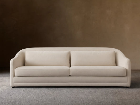 A beige Bianca Sofa with curved armrests and two large seat cushions. Made from soft performance fabric, set against a light brown wall and wooden floor.