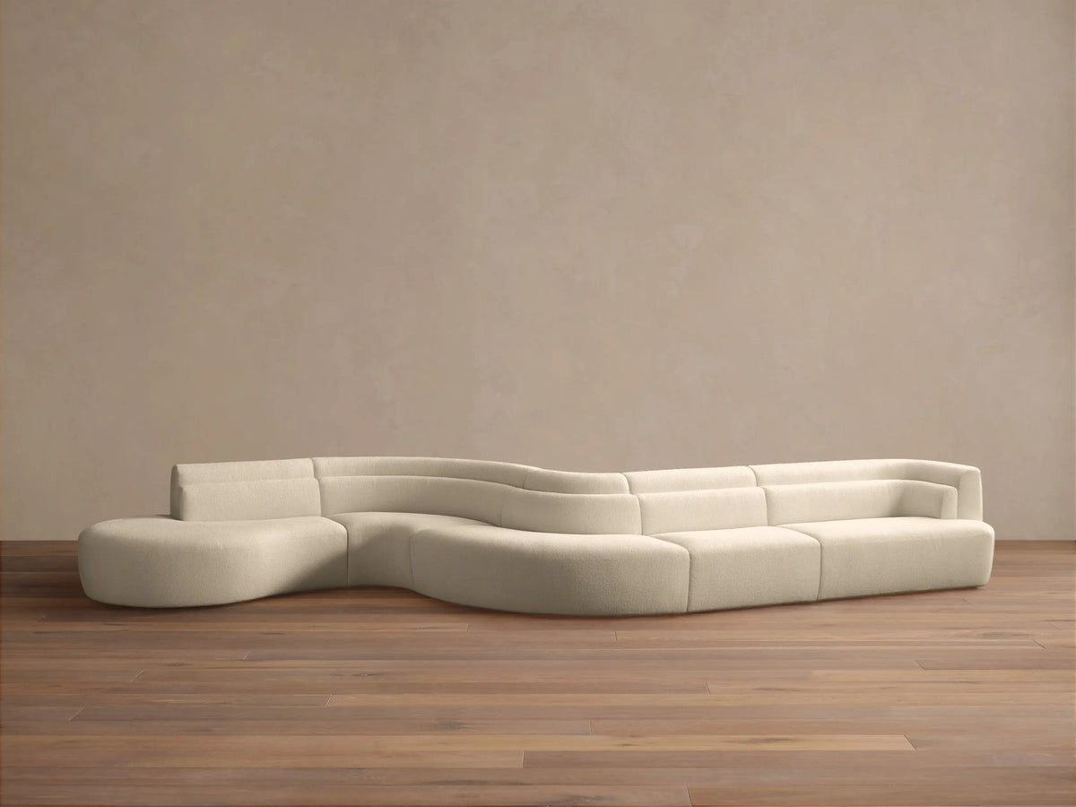 Light beige Lovina Five-Piece Sectional, a wavy, modular sofa with rounded chaise, on a wood floor against a beige wall.