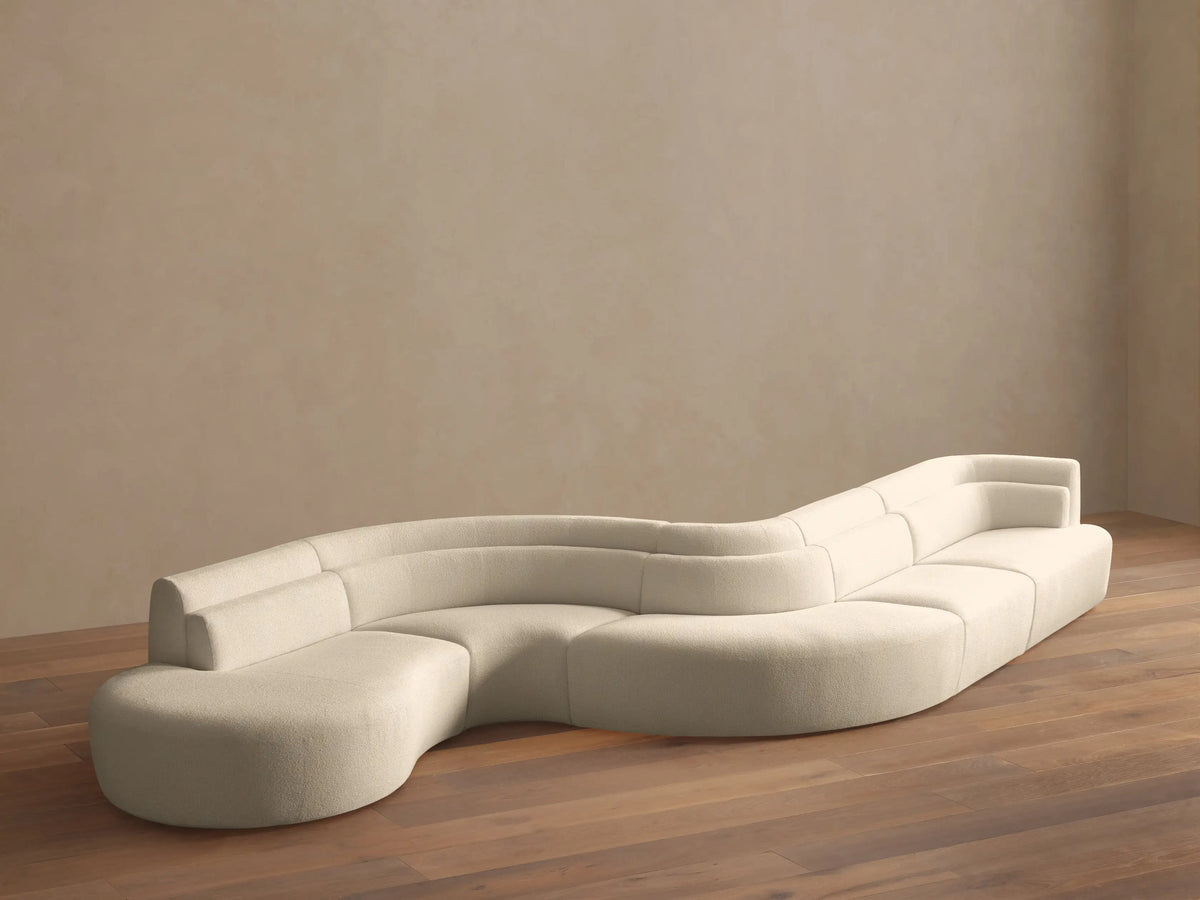 Light beige Lovina Five-Piece Sectional, a wavy, modular sofa with rounded chaise, on a wood floor against a beige wall.