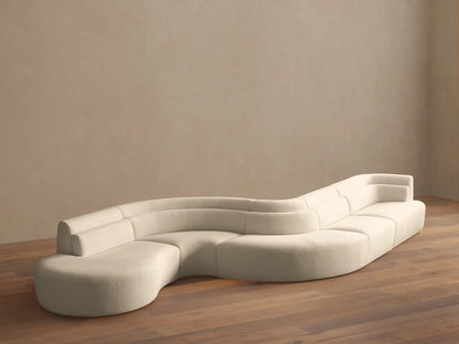 Light beige Lovina Five-Piece Sectional, a wavy, modular sofa with rounded chaise, on a wood floor against a beige wall.