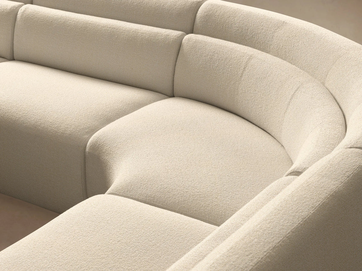 Lovina Five-Piece Corner Sectional: A large, curved light beige sofa on a warm wooden floor against a pale wall.