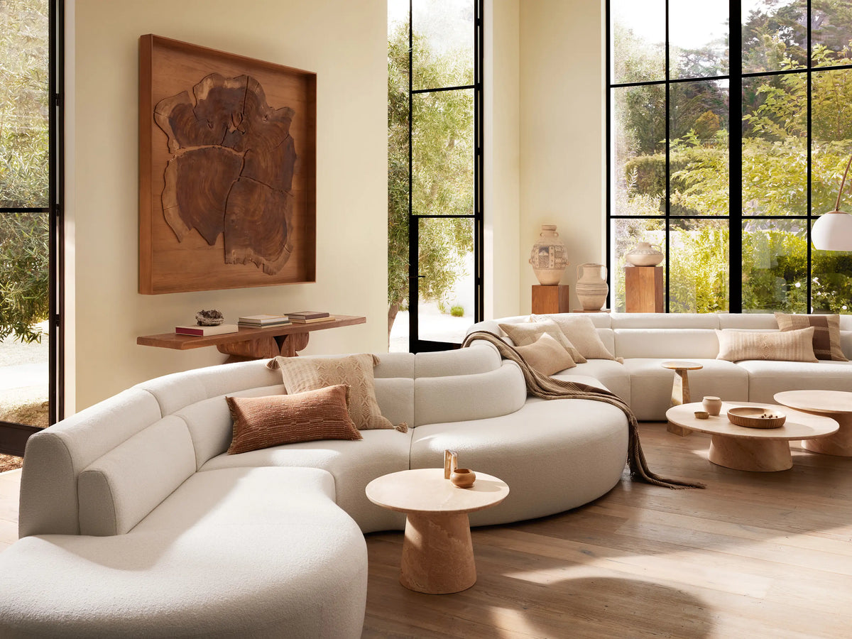 Lovina Seven-Piece Sectional, curved and upholstered in light cream textured fabric, with pillows & blanket, in a bright living room.