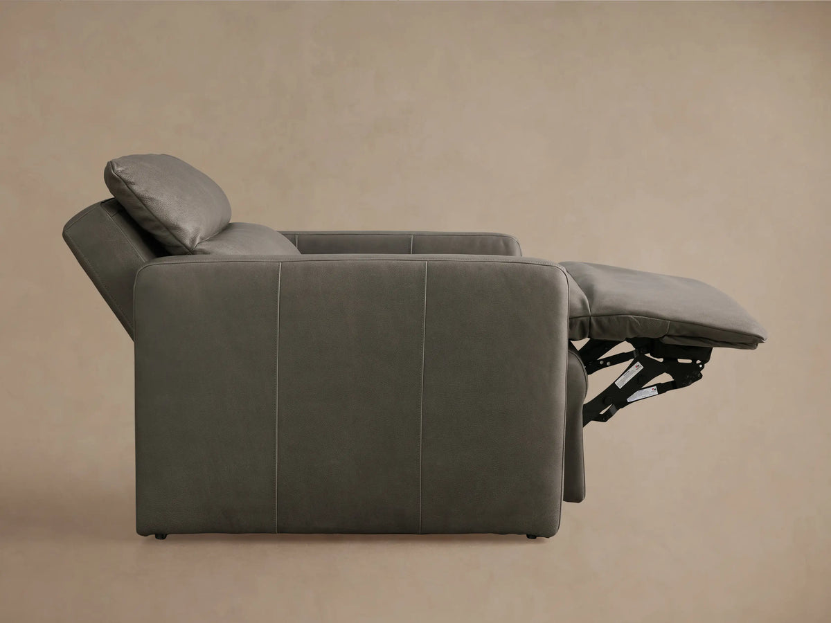 Charcoal gray Rowland Tailored Leather High-Back Motion Recliner, angled view, footrest extended, against a light brown wall.