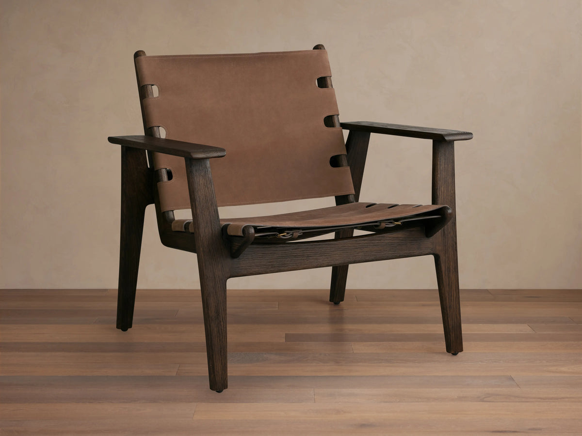 Nubuck leather chair with a sling-style seat, secured by antique brass buckles on a red oak frame, shown on wood floor.