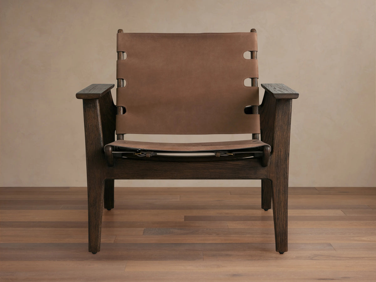 Nubuck leather chair with a sling-style seat, secured by antique brass buckles on a red oak frame, shown on wood floor.