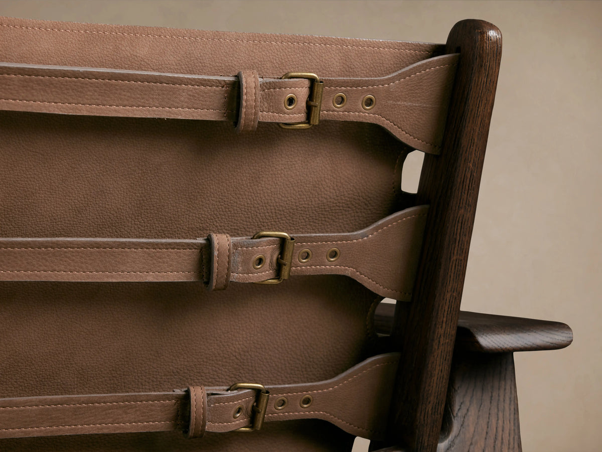 Reggie Nubuck Leather Chair with brown sling-style seat, dark oak wood frame, brass buckles, on a wooden floor.