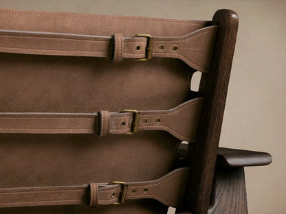 Reggie Nubuck Leather Chair with brown sling-style seat, dark oak wood frame, brass buckles, on a wooden floor.