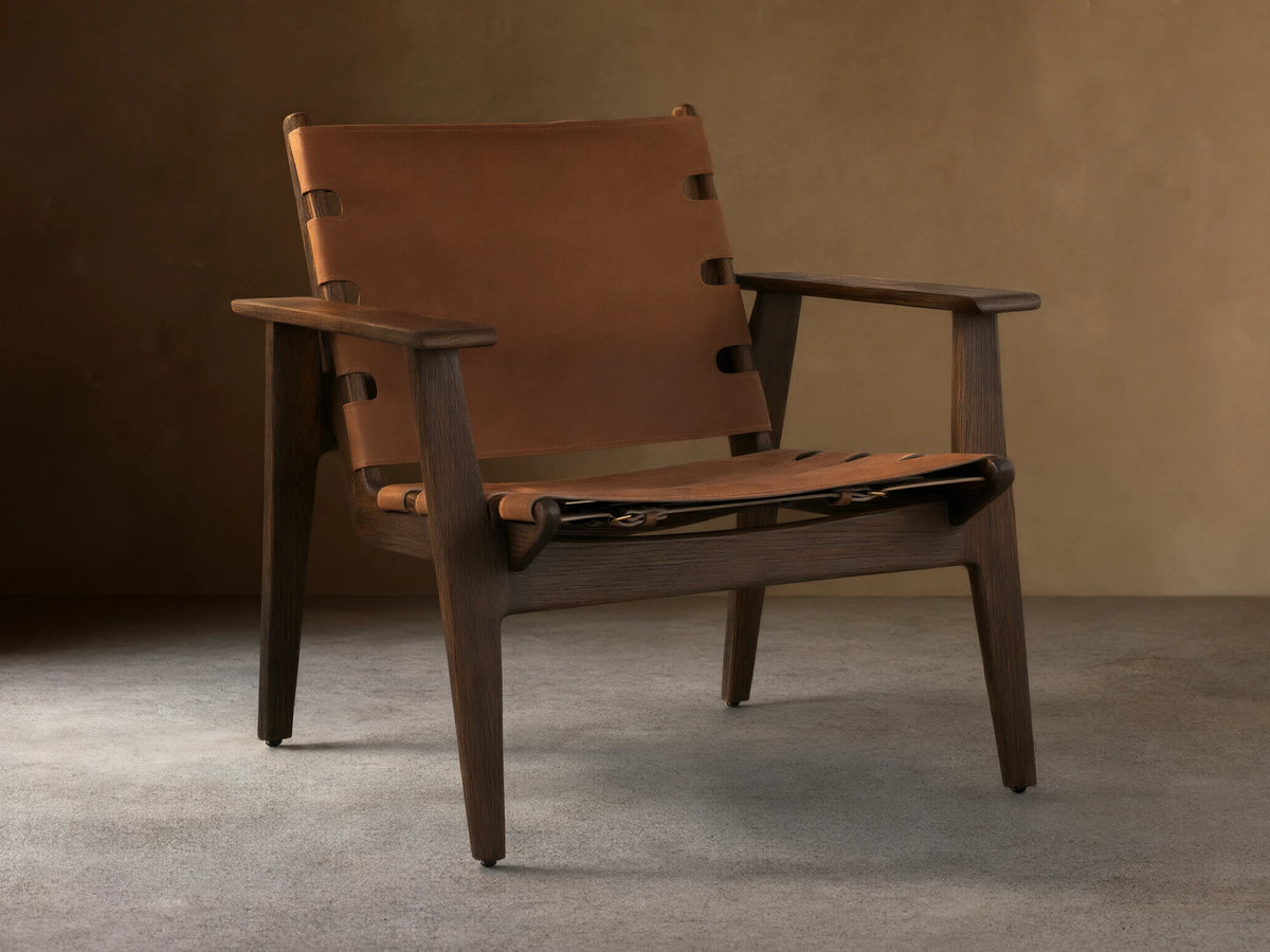 Nubuck leather chair with a sling-style seat, secured by antique brass buckles on a red oak frame, shown on wood floor.