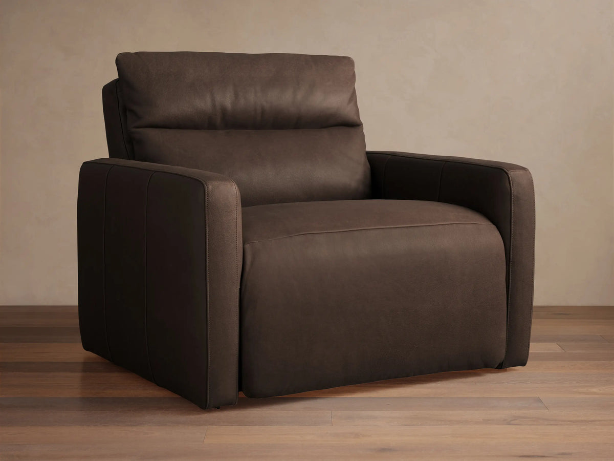 Rowland Tailored dark brown leather high-back motion recliner. Angled view, tailored design, plump cushions, on wood floor.