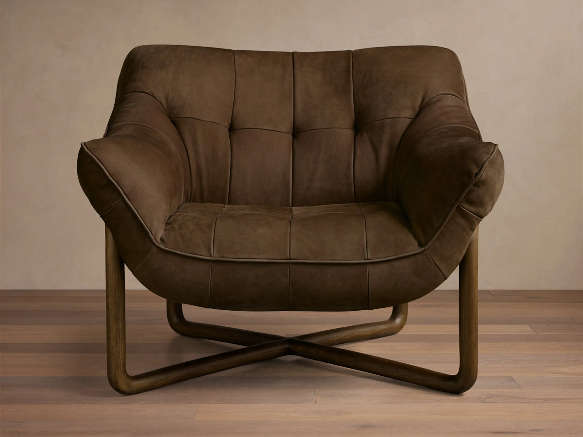 Front view of a dark brown distressed leather lounge chair with deep tufting, piping, and a curved wooden frame.