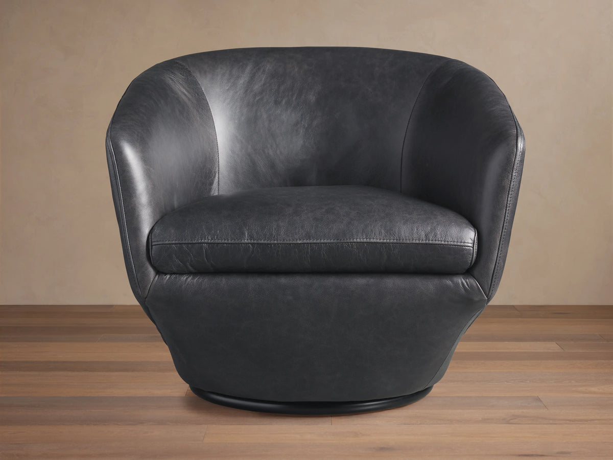 Dark gray Pelton Leather Swivel Chair, angled view, showing its curved barrel shape and round black swivel base on a wood floor.