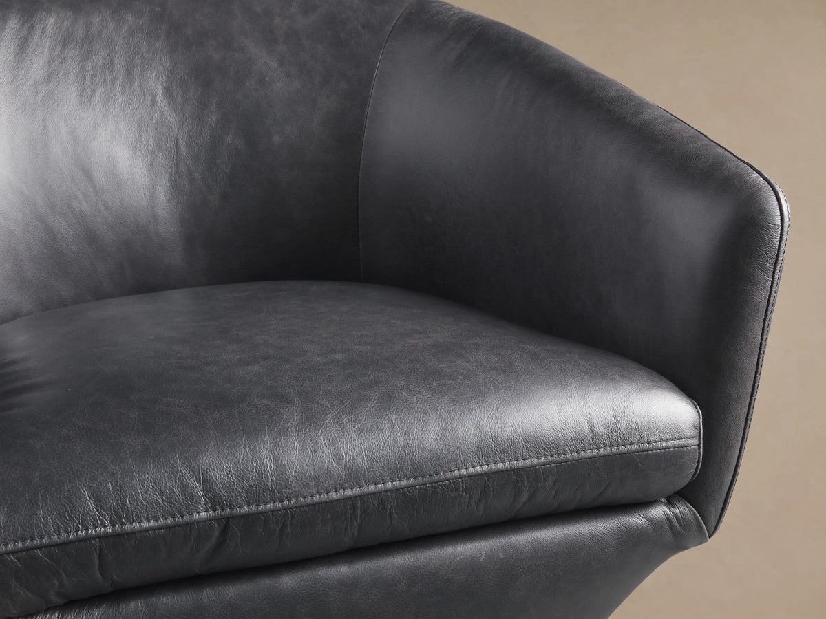 Pelton Leather Swivel Chair. Dark gray leather, curved back with vertical stitching, shown from the side-back view on a beige background.