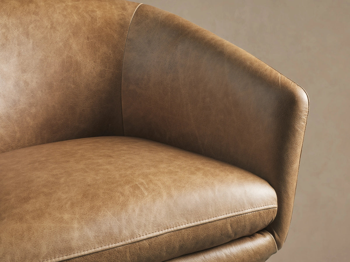 Close-up of the Pelton Leather Swivel Chair, showing a warm, distressed brown leather seat and curved armrest with fine stitching.