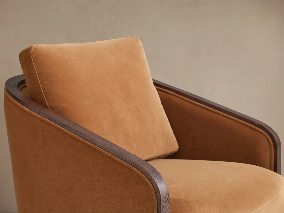 Pomona Swivel Chair with curved back in warm camel fabric, dark oak wood trim, and circular swivel base, angled from rear.