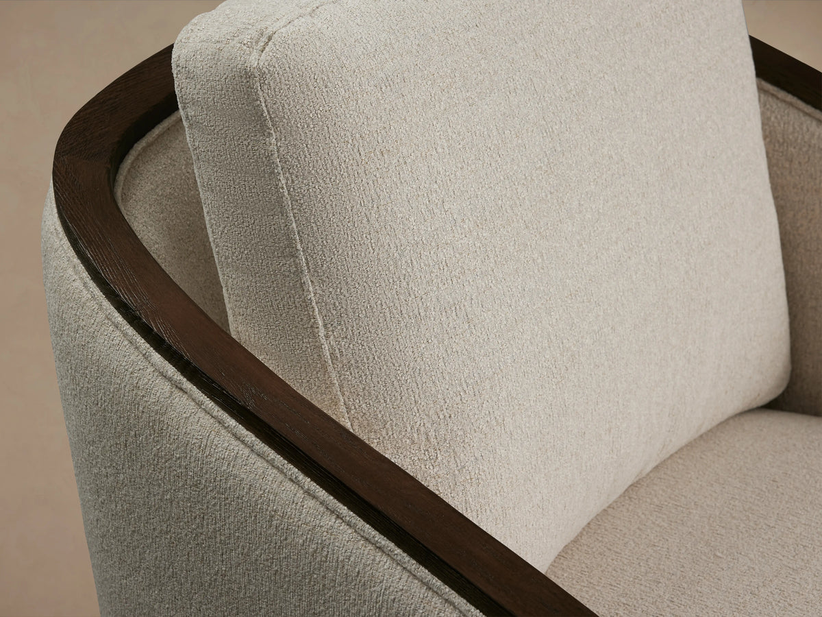 Pomona Swivel Chair with light beige fabric, dark wood exposed arms, and base, on wooden floor against a neutral wall.