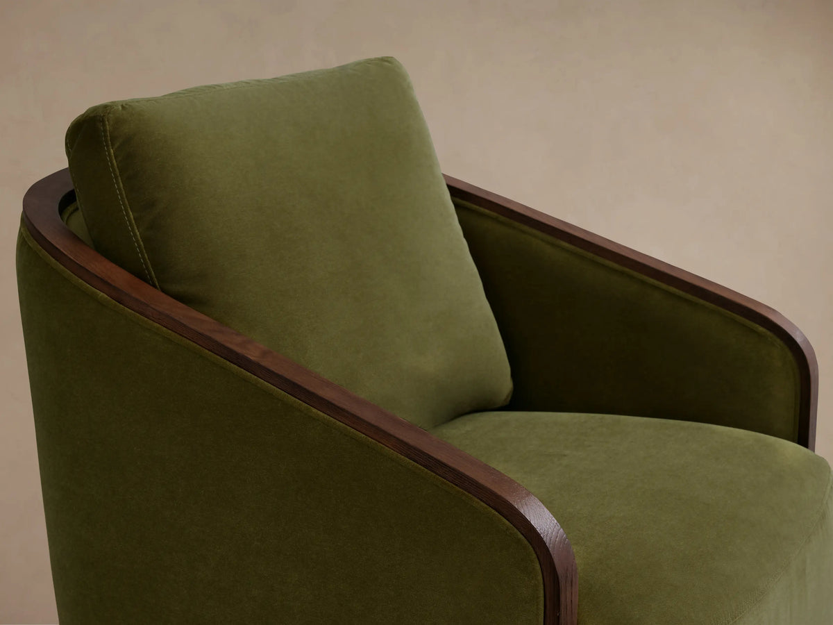 Pomona Swivel Chair with olive green fabric, curved exposed oak arms, and a round base, on a wooden floor background.