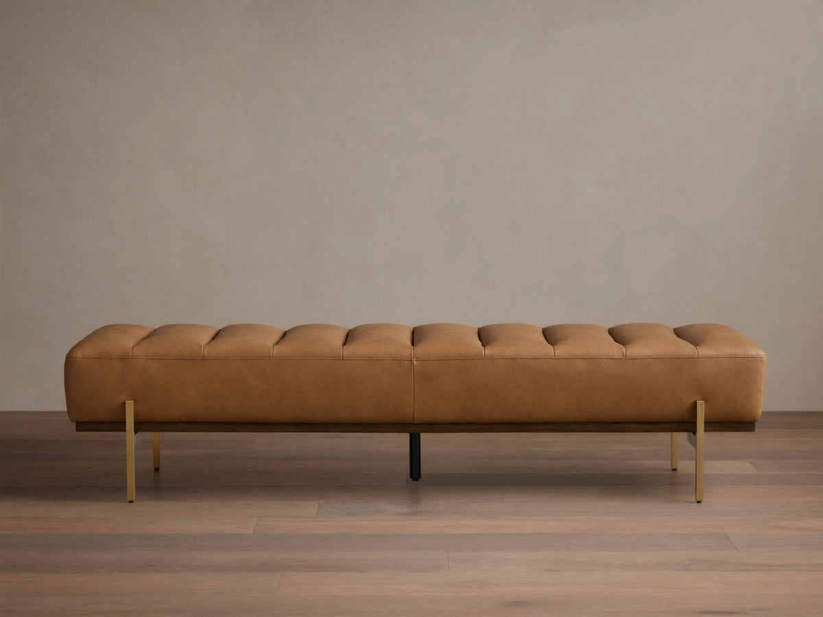 Front view of a camel leather tufted bench with a dark wood base and slender gold-toned metal legs.