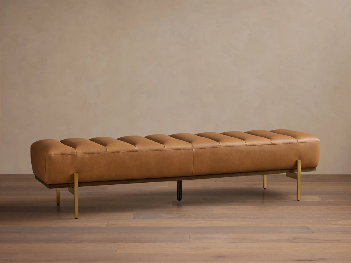 Front view of a camel leather tufted bench with a dark wood base and slender gold-toned metal legs.