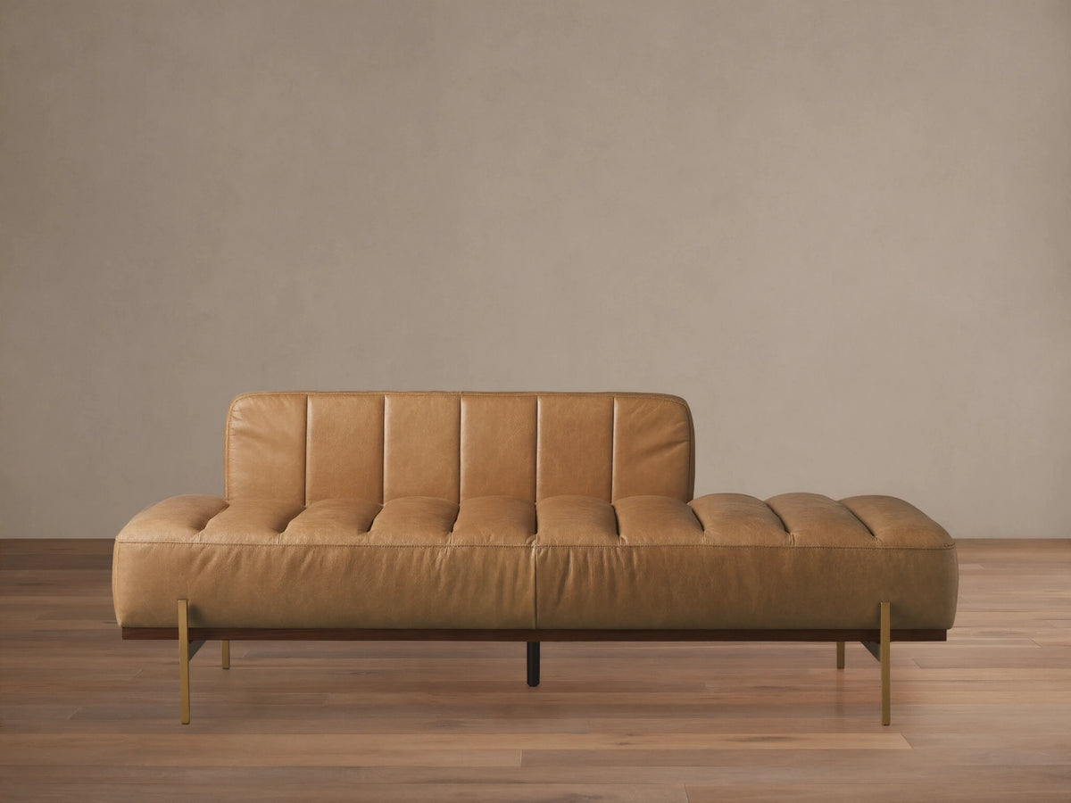 Lansing Leather Daybed with tan channel-tufted leather, low backrest on left, dark wood base, and gold legs on wood floor.