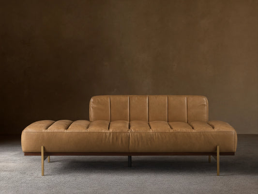 Lansing Leather Daybed with tan channel-tufted leather, low backrest on left, dark wood base, and gold legs on wood floor.