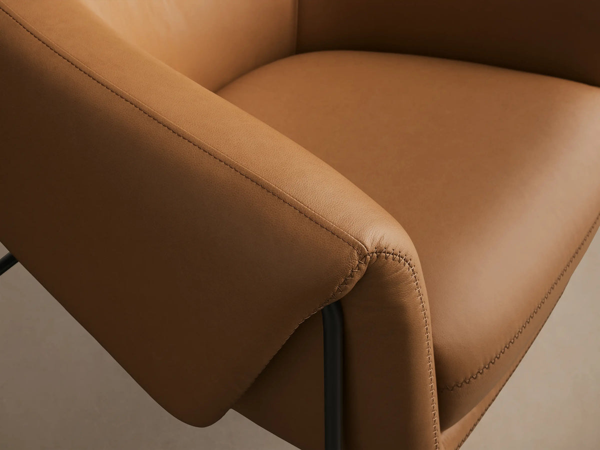 Front view of a Caramel Linus Leather Chair with a curved back, prominent stitching, and sleek black metal legs.
